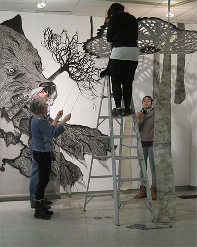 Students install large wall drawing using ladder and cut materials in gallery