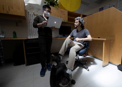 Person showing a laptop screen to another seated person wearing a sensor cap in a lab workspace.