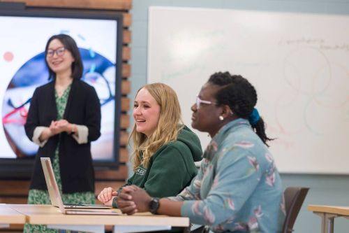 Classroom with two students at a table and an instructor leading a lesson near a screen and whiteboard.