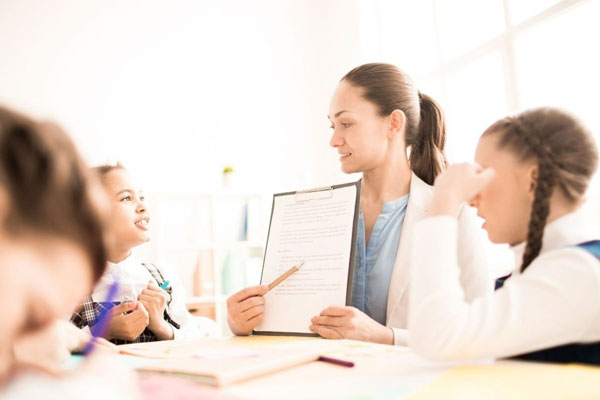 A teacher holds a clipboard and points to a worksheet while young children sit around the table during a classroom activity.