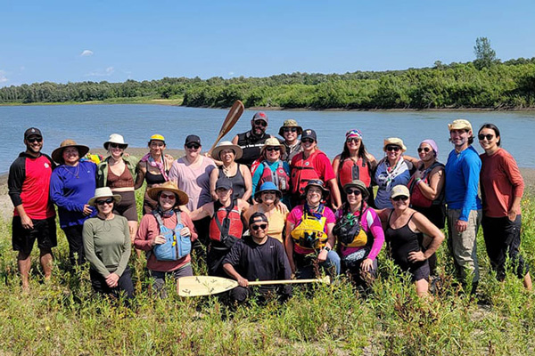 A large group of people in life jackets and outdoor clothing pose together on a grassy riverbank, with paddles resting in front of them. A wide river and tree‑lined shoreline stretch behind the group under a clear blue sky.