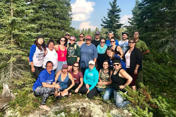A large group of people standing and kneeling together on a rocky, forested area, surrounded by evergreens under a bright sky. They appear to be on an outdoor trip or group retreat, posing for a group photo in nature.