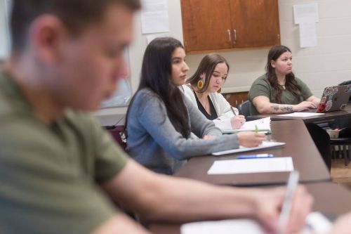 A group of students sit at tables in a classroom, writing notes during a discussion, with notebooks and laptops in front of them.