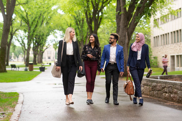 Group of business‑dressed students walking together on a tree‑lined campus path.