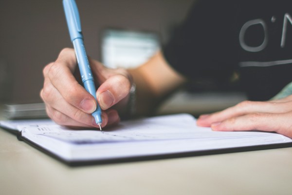 Close-up of a hand writing in a notebook with a blue pen on a desk.