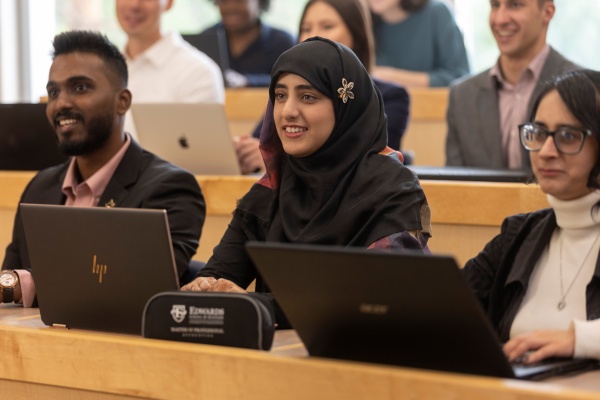 Students seated in a classroom setting with wooden desks, using laptops. A nameplate on one desk reads “Edwards School of Business.” The background shows more students and laptops, indicating a lecture or group learning environment.