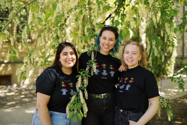 Three people in black shirts stand together outdoors beneath green leaves and hanging blossoms.