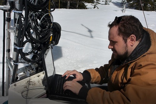 Person uses a laptop connected to field instruments in a snowy outdoor hydrology research site.