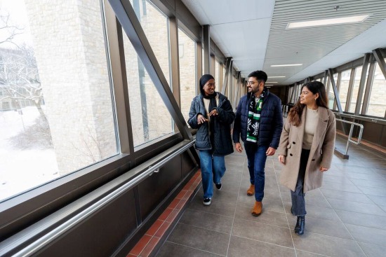 Three people walk through a glass‑walled campus corridor with windows and snow outside.