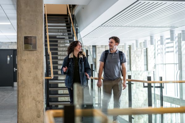 Two students walking together in modern campus hallway by stairs and windows.