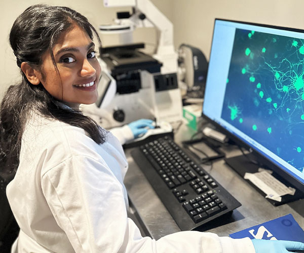 Person wearing a lab coat and gloves works at a laboratory bench beside a microscope, viewing fluorescent cell images on a computer monitor.