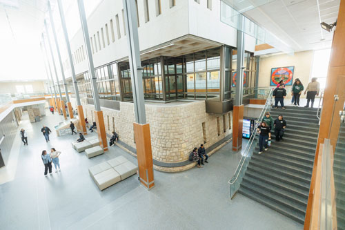A bright, modern academic building atrium with high ceilings, large windows, and multiple levels. Students gather on seating areas and stairs, walk through open corridors, and interact in a shared central space.