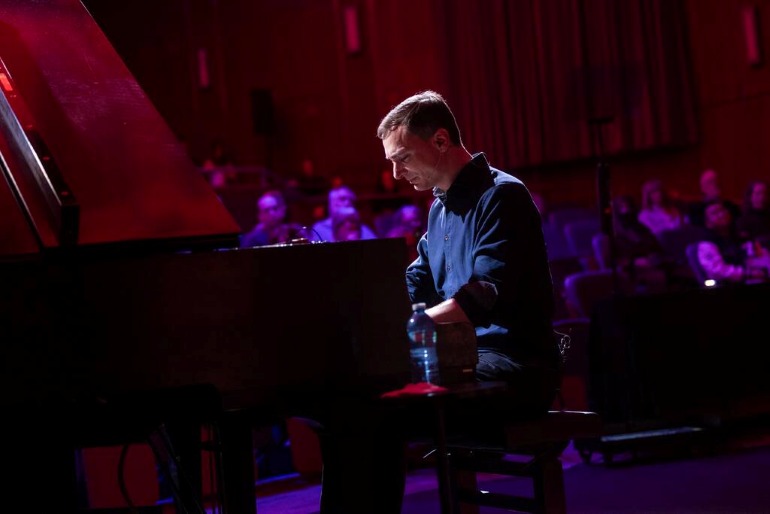Person playing a grand piano on a dimly lit stage with an audience seated behind.