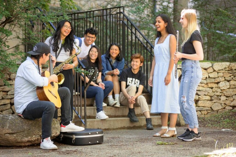 Small group of people making music outdoors with guitar, saxophone, flute, and voices.