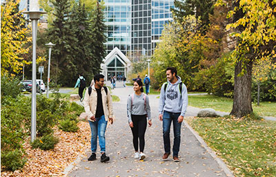 Three students walk together along a tree‑lined campus pathway during autumn, surrounded by colourful leaves and greenery, with a modern glass building visible in the background.