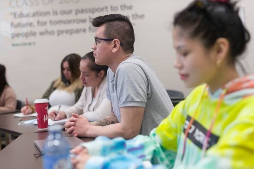 Students seated at a classroom table, listening and taking notes with notebooks, laptops, and coffee cups visible in a collaborative learning setting.