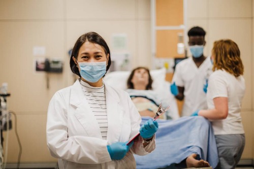 Healthcare training scenario with a person in a lab coat holding a clipboard in the foreground and two individuals attending to a patient on a hospital bed in the background