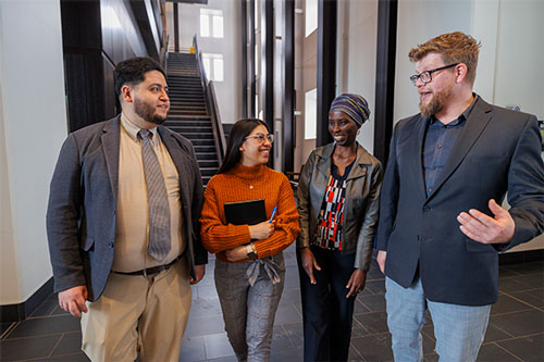 Four people stand together in an indoor building atrium, engaged in conversation near a staircase. They appear to be in a professional or academic setting, with two holding notebooks and all dressed in business or business‑casual clothing.