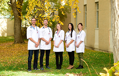 Five people wearing white lab coats stand in a row outdoors on a grassy area with autumn leaves. They are smiling, with a building wall and trees with yellow foliage in the background.