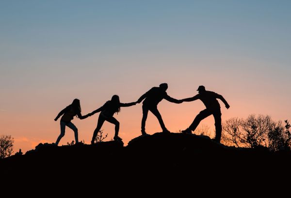 Four people hold hands, helping each other climb a ridge at sunset.